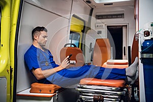 Male paramedic in a blue uniform resting and browsing his phone in the ambulance car