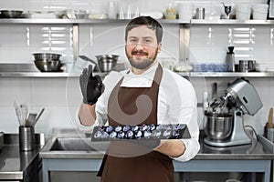 Male pastry chef in a professional kitchen holds handmade chocolates in his hands and shows the ok sign