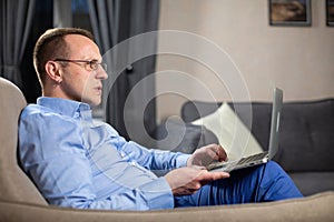 Male person sits in armchair and looks at screen of laptop