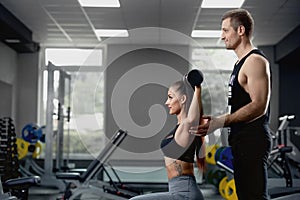 Male personal trainer helping woman working with heavy dumbbells at gym.