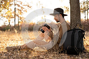 Male photographer in the autumn forest sitting under a tree looking at the results of filming on the screen of a digital