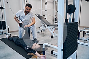 Male physical therapist bends female knee with rehab equipment strap in therapeutic office