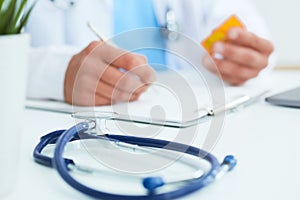 Male physician medicine doctor or pharmacist sitting at worktable, holding jar or bottle of pills in hand and writing