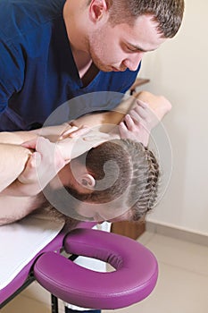 A male physiotherapist does a power stretching for a male patient to a young bearded hipster in a medical massage room