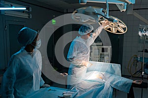 Male plastic surgeon in surgical uniforms and masks adjusting light before operation in dark operating theatre. Surgery