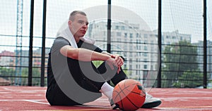 Male player sitting on basketball sports ground with ball. Concentrated sportsman looking at camera during training.