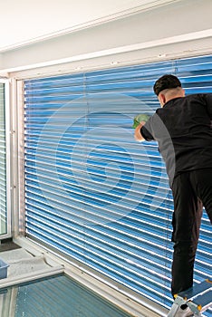 Male professional cleaner cleaning blue blinds on a large window front with micro fiber cloths