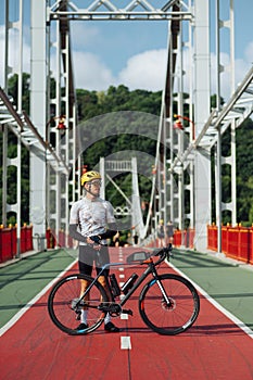 Male professional cyclist stands with a bicycle on the bridge and looks away with a serious face
