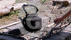 Male Puts Dry Branches in a Fire with a Kettle Boiling over the Grate in Smoke