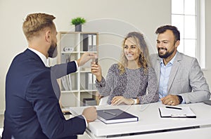 Male real estate agent at meeting in office hands over keys to his new home to joyful young couple.
