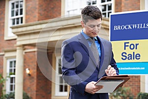 Male Realtor Standing Making Notes Outside Residential Property With For Sale Sign photo