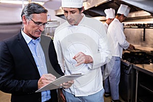 Male restaurant manager writing on clipboard while interacting to head chef