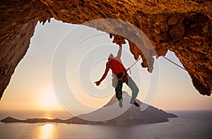 Male rock climber hanging on cliff with one hand at sunset.