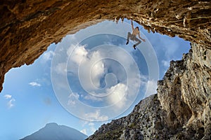 Male rock climber hanging with one hand on on a cliff against sky