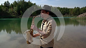 Male saxophonist is playing melody on shore of picturesque forest lake at summer day