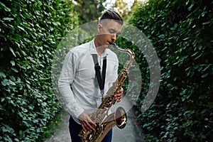 Male saxophonist plays on the alley in summer park