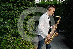 Male saxophonist plays on the alley in summer park