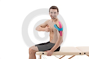 Male shoulder with physio tape,  on a white background. Male model sitting on a portable massage table