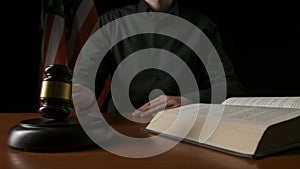 Male sitting at the desk with wooden hammer and law book, USA flag at the background. Man judge in the court room