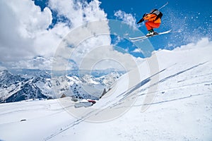 A male skier in an orange suit flies in the air after jumping from a snow sweep high in the Caucasian mountains on a