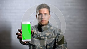Male soldier in uniform showing smartphone with green screen at camera, app
