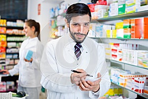 Male specialist is attentively stocktaking medicines with notebook near shelves in pharmacy.