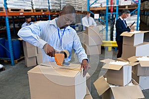 Male staff packing cardboard box with tape gun dispenser in warehouse