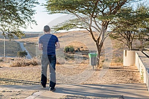 Male standing near the trees and looking at the road and the hills on a sunny day