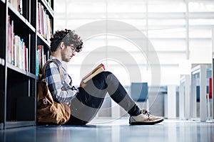 Male student in library, looking for books, preparing for final exam. Reading and sitting on floor.