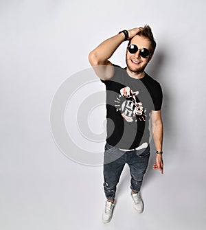 Male in sunglasses, black t-shirt, bracelets, jeans and sneakers. Smiling, showing thumbs up posing isolated on white. Full length