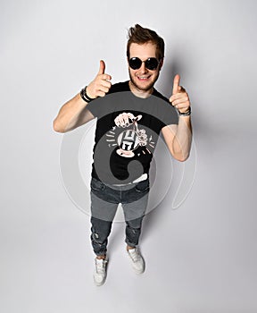 Male in sunglasses, black t-shirt, bracelets, jeans and sneakers. Smiling, showing thumbs up posing isolated on white. Full length