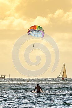 Male surfer sitting on his board looking at a rainbow parasailing parachute in Hawaii