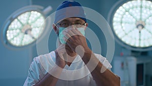 A male surgeon puts on his medical mask while standing in the middle of the surgery room, he is wearing a dark blue hair