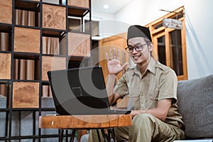 male teacher in civil servant uniform and wearing a cap waving in front of a laptop computer