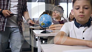 Male teacher of elementary school walking between desks puts computers tablet on desk pupils for learning