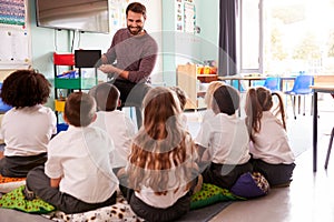 Male Teacher Holding Digital Tablet Teaches Group Of Uniformed Elementary Pupils In School Classroom