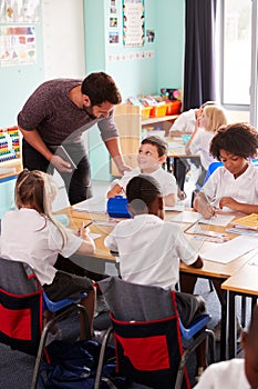 Male Teacher Holding Digital Tablet Teaches Group Of Uniformed Elementary Pupils In School Classroom