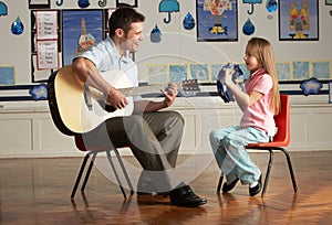 Male Teacher Playing Guitar With Pupil In Classroo