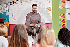 Male Teacher Reading Story To Group Of Elementary Pupils In School Classroom