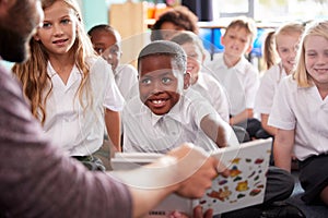 Male Teacher Reading Story To Group Of Elementary Pupils Wearing Uniform In School Classroom photo