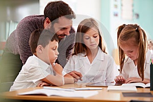 Male Teacher With Three Elementary School Pupils Wearing Uniform Using Digital Tablet At Desk