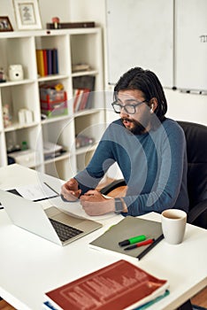 Male teacher wearing eyeglasses sitting at his workplace and teaching HTML CSS online, looking at webcam and talking photo
