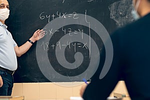 Male teacher wearing a face mask and explaining equations on a blackboard to the students