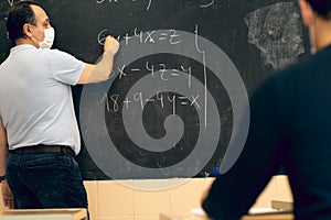 Male teacher wearing a face mask and writing equations on a blackboard