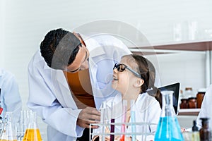 A male teacher in white lab coat ask a little Asian girl how about doing experiment. A girl with glasses explain how to process