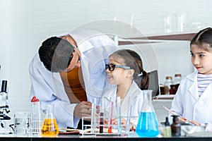 A male teacher in white lab coat ask two little Asian girls how about doing experiment. A girl with glasses explain how to process