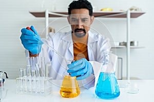 A male teacher in white lab coat with blue rubber gloves with many laboratory tools on shelves and table. Using pipette right hand