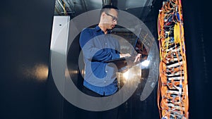 A male technician is standing in a server room and operating his laptop