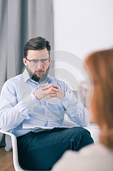 Male therapist sitting on a chair with joined hands and listening to his patient