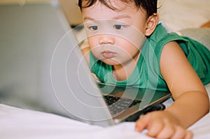 A male toddler is playing with a laptop while on the bed